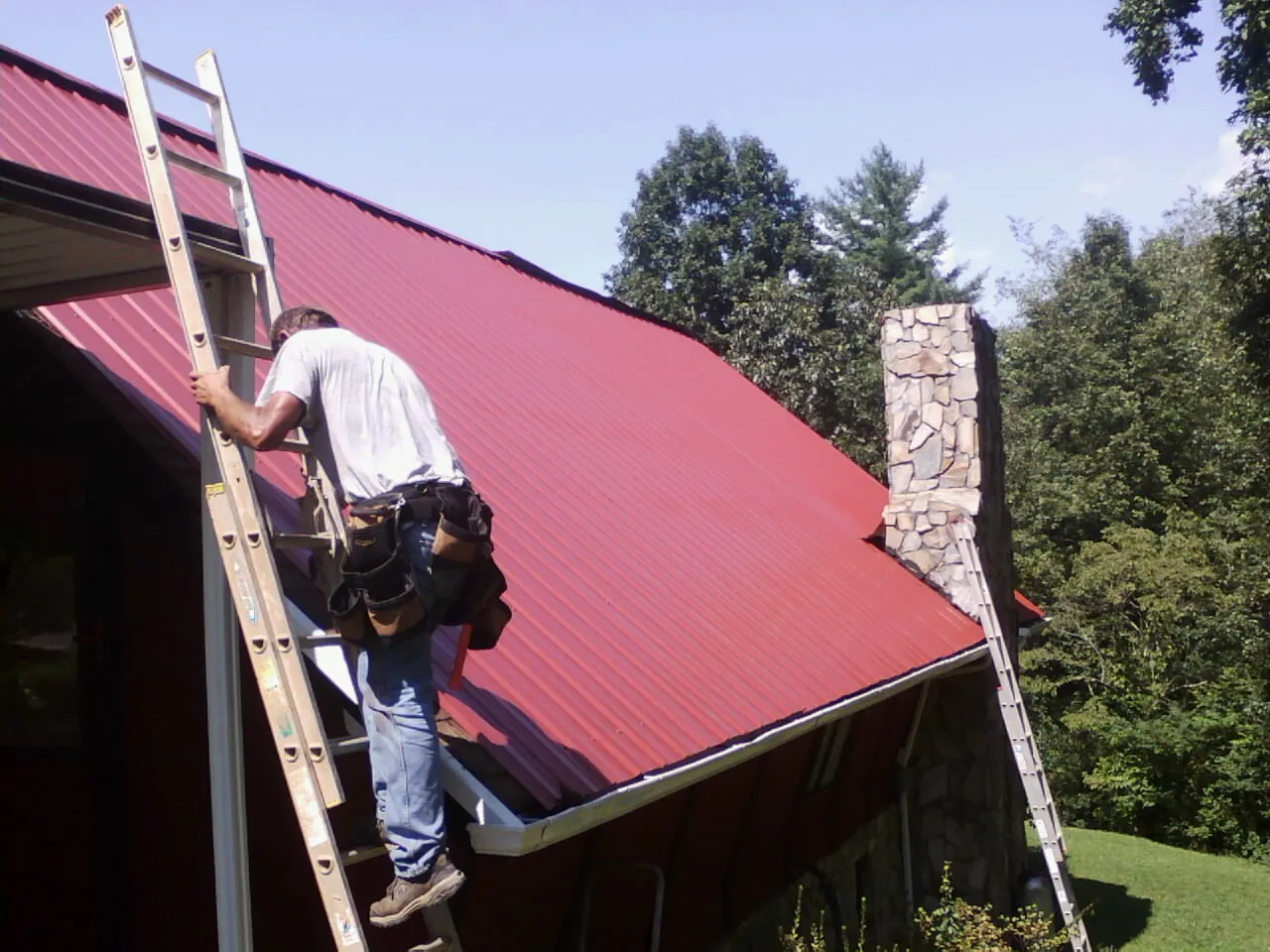 Roofer on a ladder beside a red metal roof and stone chimney — Soffit & Fascia Repair by Roofing Brothers in Boxborough